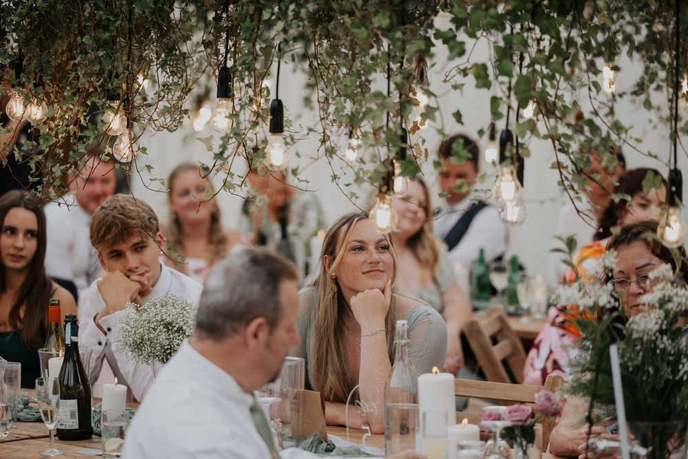 Wedding guests smile and listen attentively at reception table adorned with hanging Edison bulb lights.