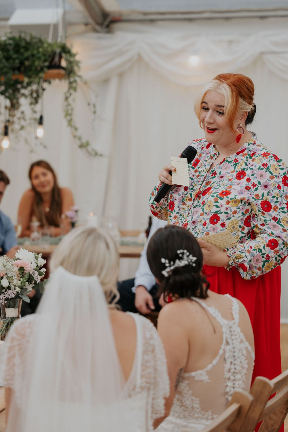 Wedding speaker in floral blouse and red skirt addresses two brides at decorated reception table.