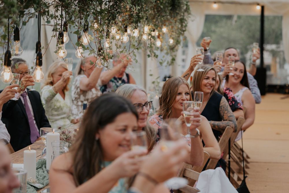 Wedding guests raising glasses in a toast under hanging Edison bulb lights and greenery at an elegant reception.
