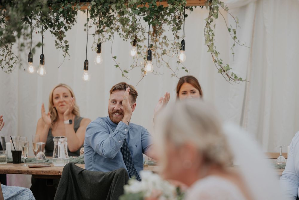 Wedding guests laughing and clapping at a reception under hanging Edison bulbs and lush greenery decor.