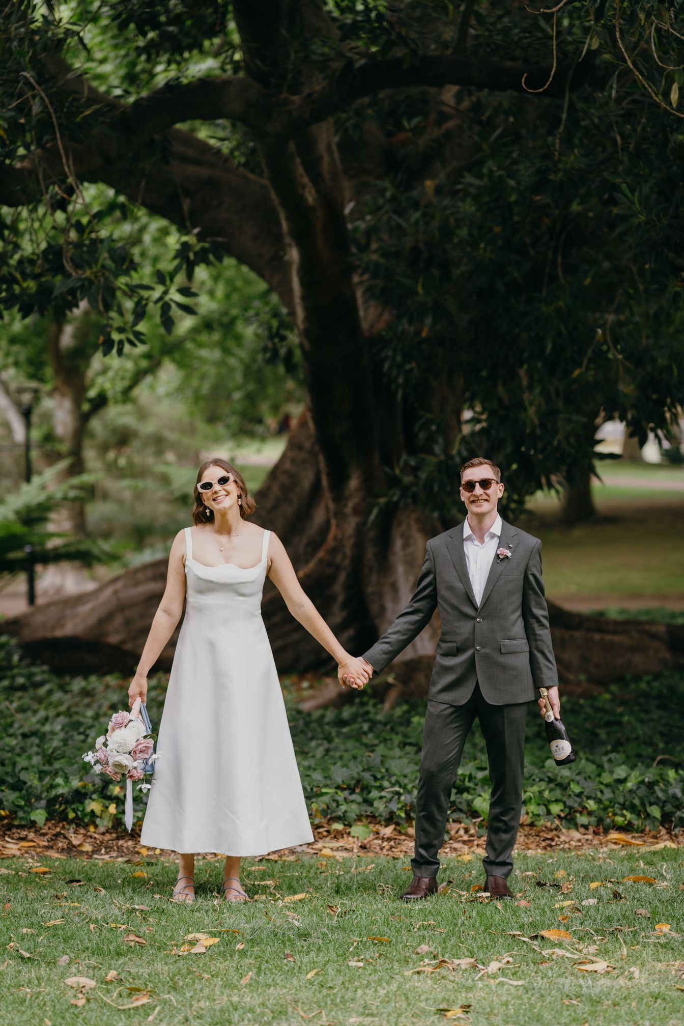 Couple holding hands while walking through a lush garden path with sunlight filtering through trees.