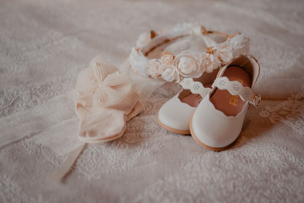 Delicate white baby shoes and accessories arranged on a soft fabric surface.
