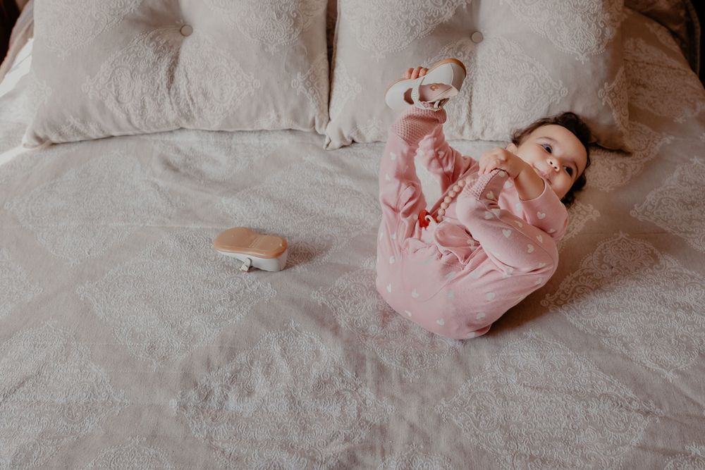 A baby in a pink outfit lies on white bedding during a lifestyle photography session.