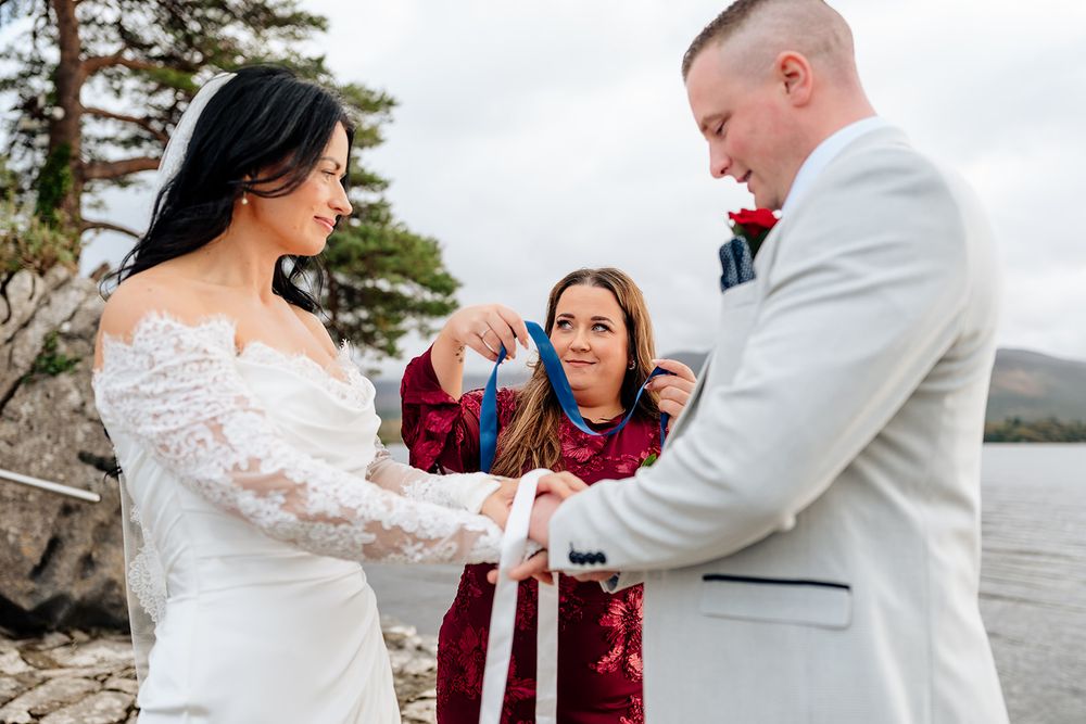 A couple shares an intimate moment during their wedding ceremony by a scenic waterfront with pine trees in the background.