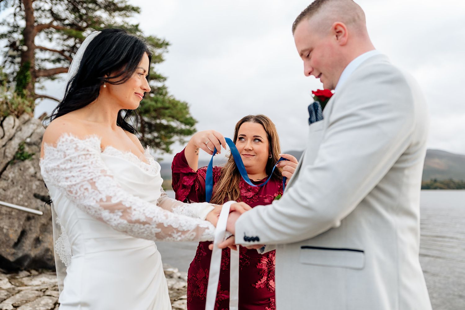 A couple shares an intimate moment during their wedding ceremony by a scenic waterfront with pine trees in the background.