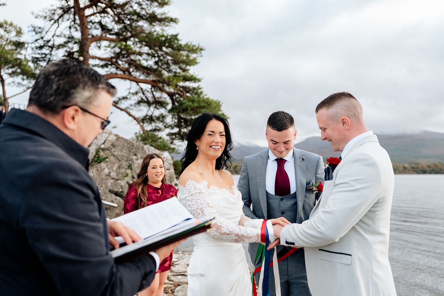 A couple signs marriage documents during an outdoor lakeside wedding ceremony with mountains in the background.