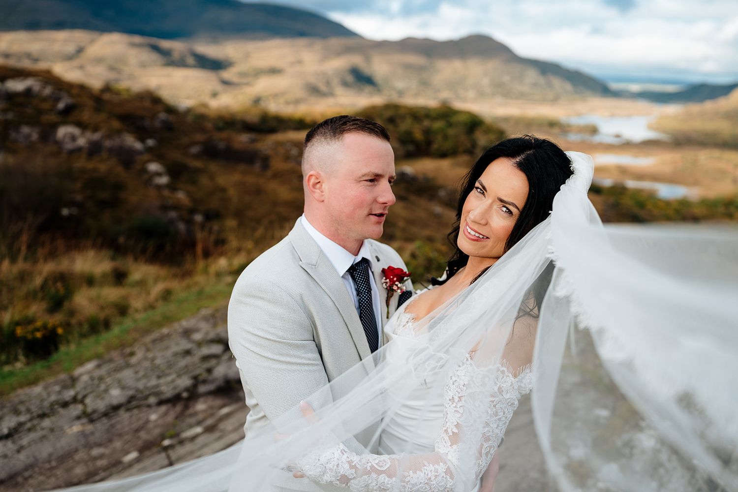 A couple in wedding attire embraces against a scenic mountain landscape with a lake in the background.