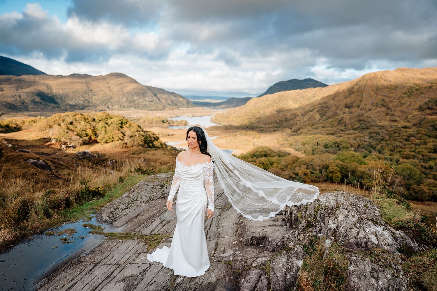A bride in a flowing white dress stands gracefully on a mountain ridge overlooking a dramatic Scottish Highland landscape.