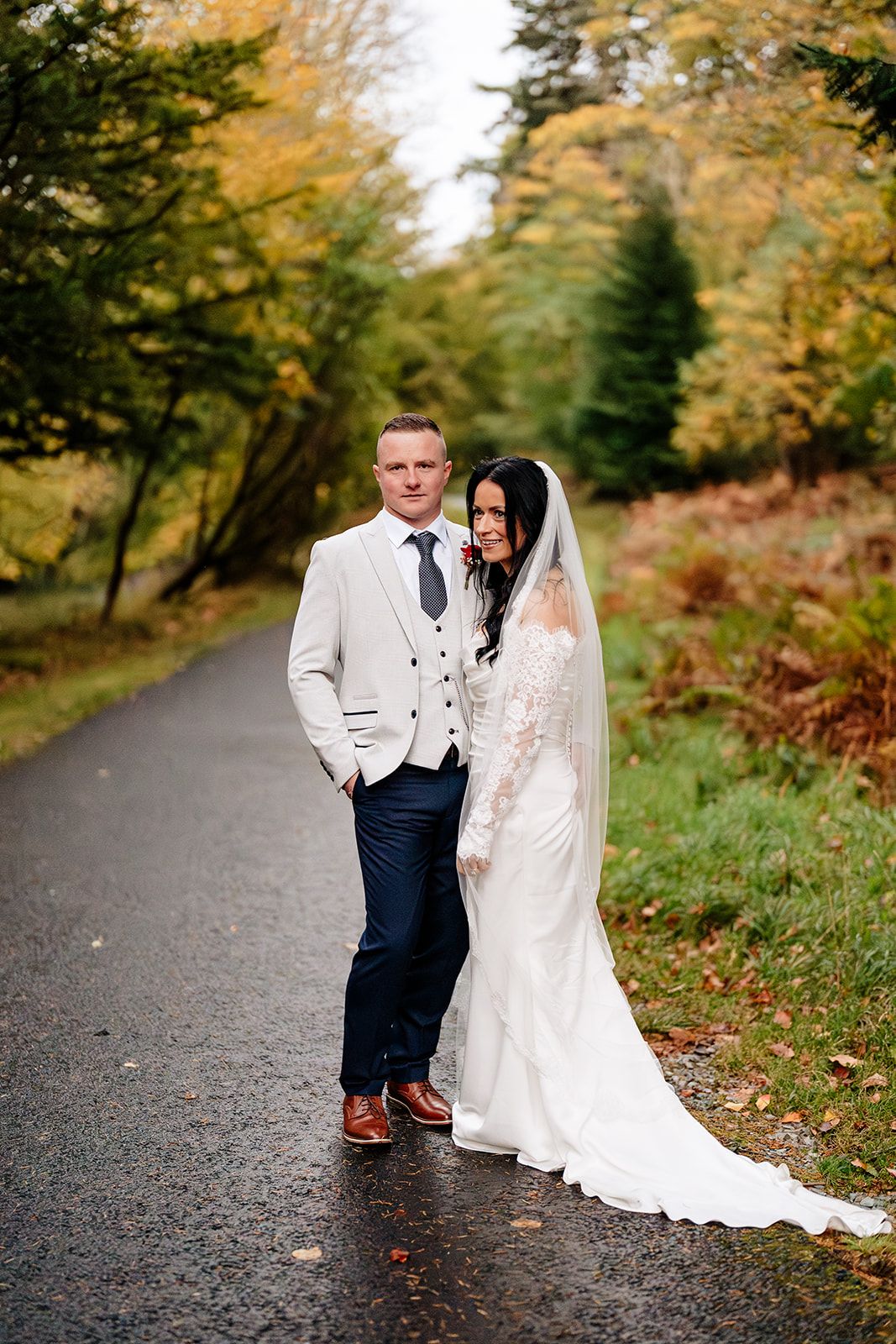 A couple poses on a wet path surrounded by vibrant autumn foliage in their elegant wedding attire.
