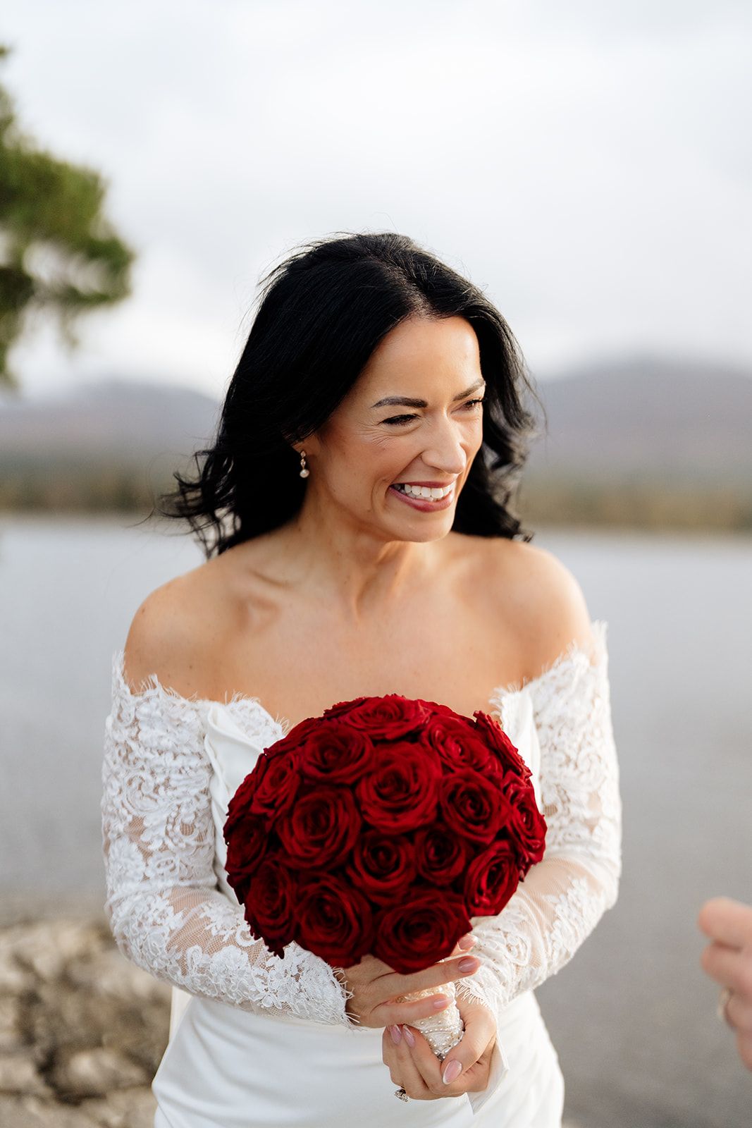 A bride in a white lace dress holds a vibrant bouquet of deep red roses against a lake backdrop.