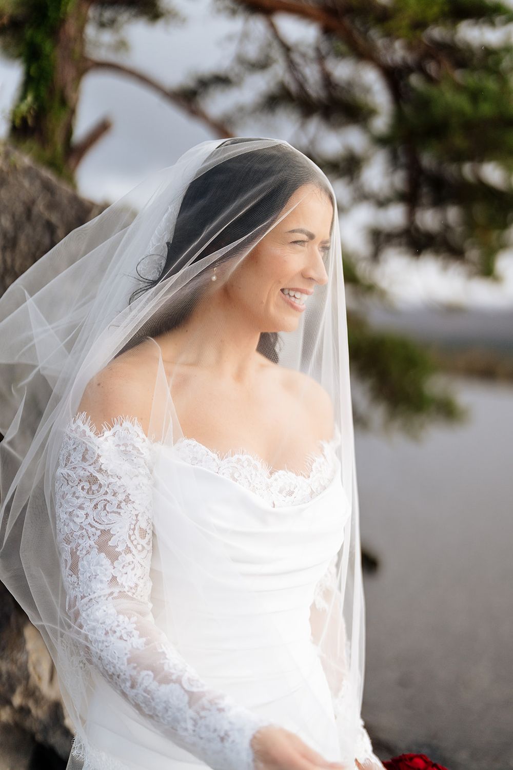 Close-up details of a delicate white lace wedding dress and veil against a soft outdoor background.
