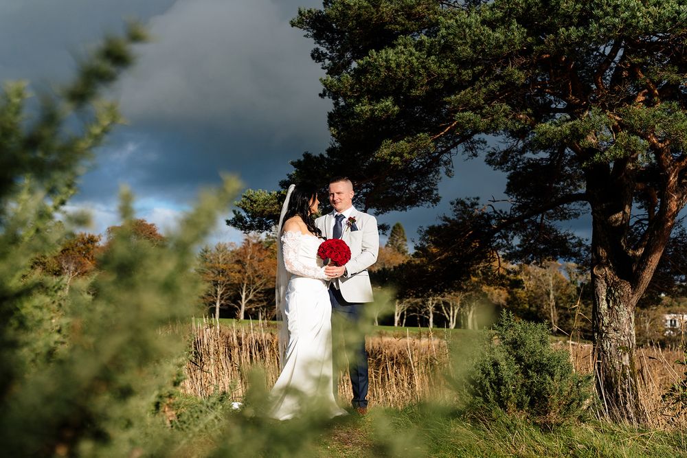 A couple in white wedding attire embraces while standing in a scenic outdoor setting with trees and natural light.