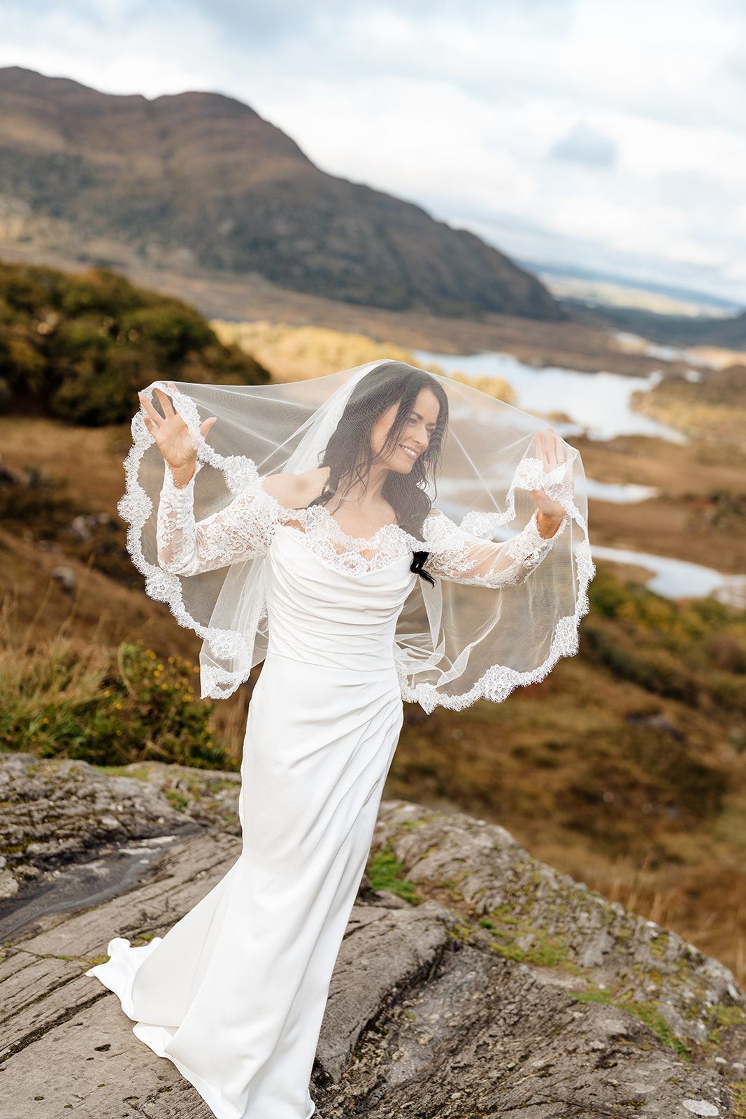 A flowing white wedding dress with a lace veil billows in the wind against the Irish mountain landscape by Sarah Kate Photography.