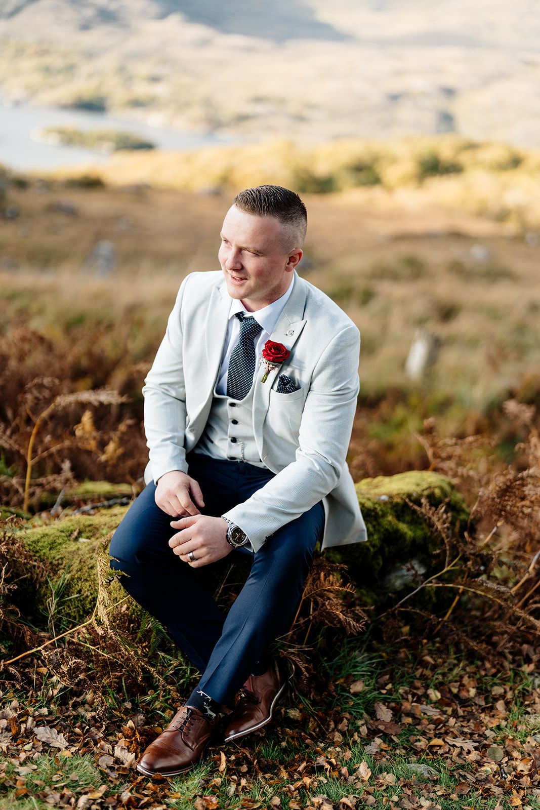 A groom in a light gray suit sits thoughtfully among autumn ferns during an outdoor wedding photo session.