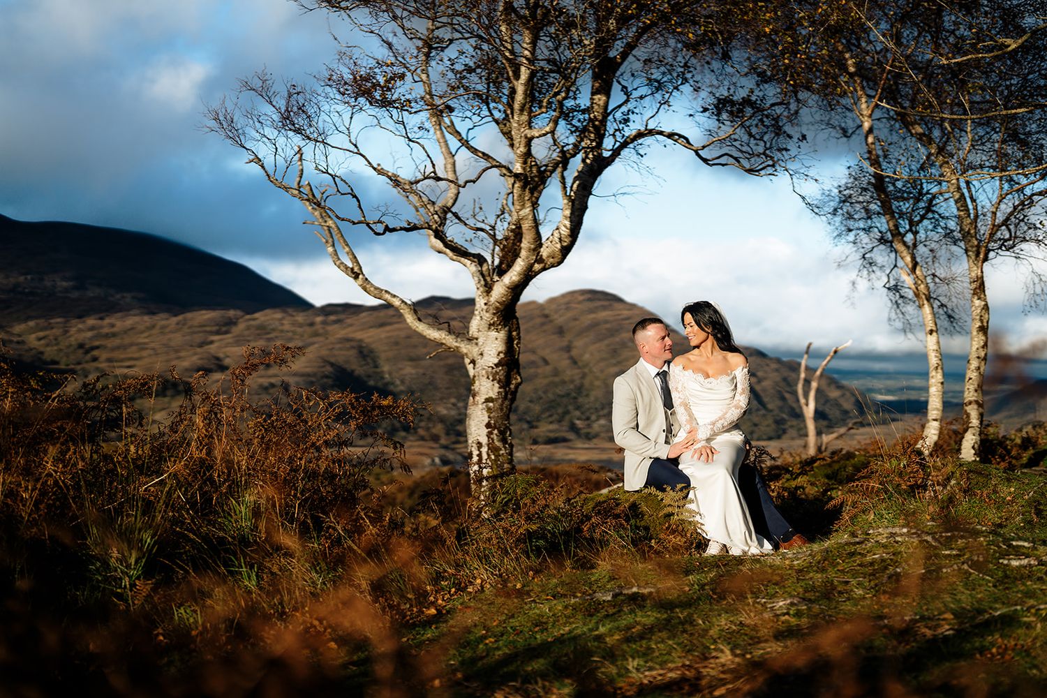 Couple in white outfits embracing against scenic mountain landscape with birch trees at sunset.