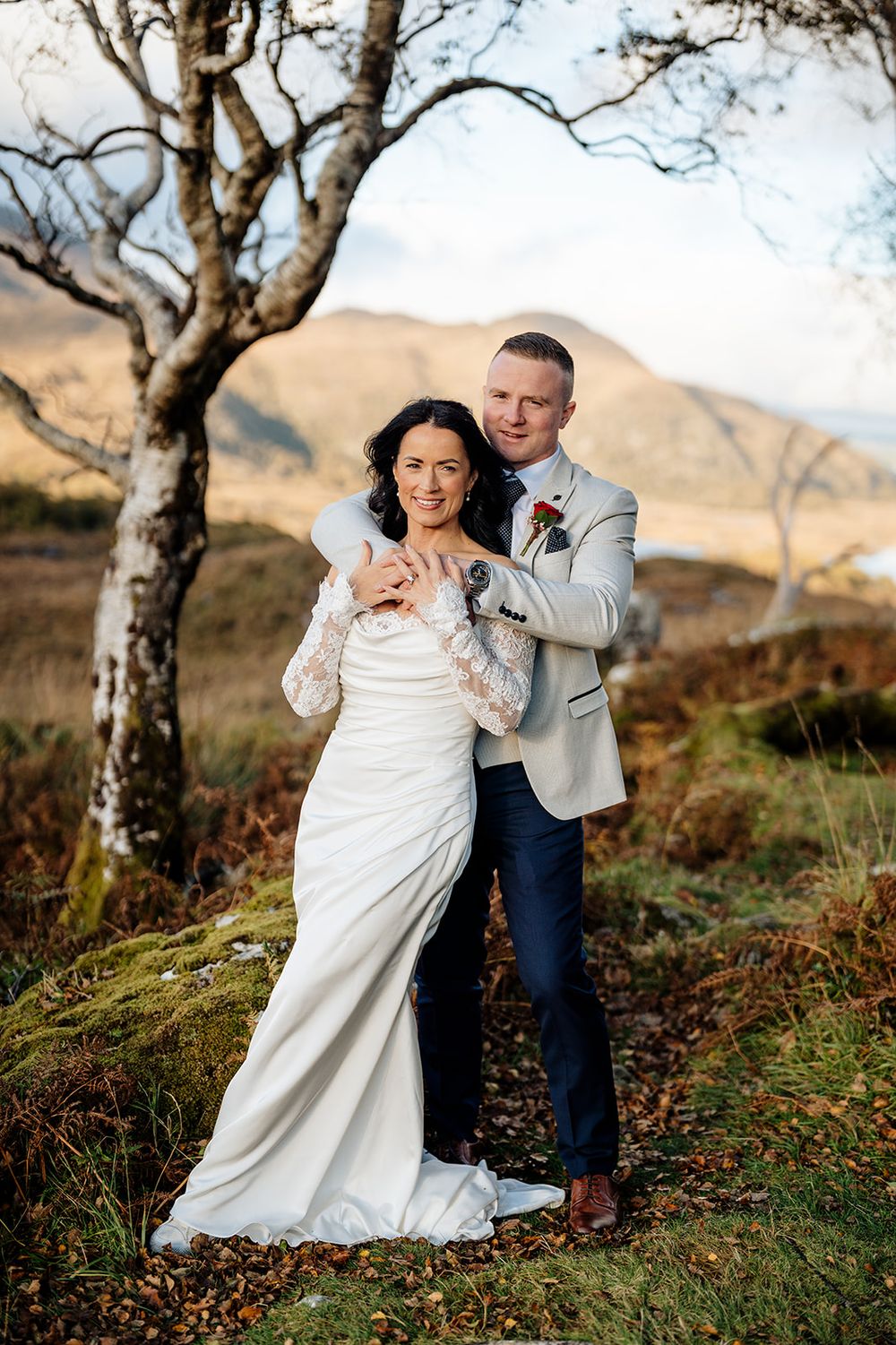 A couple in wedding attire playfully poses together on a hillside with bare trees and mountains in the background.