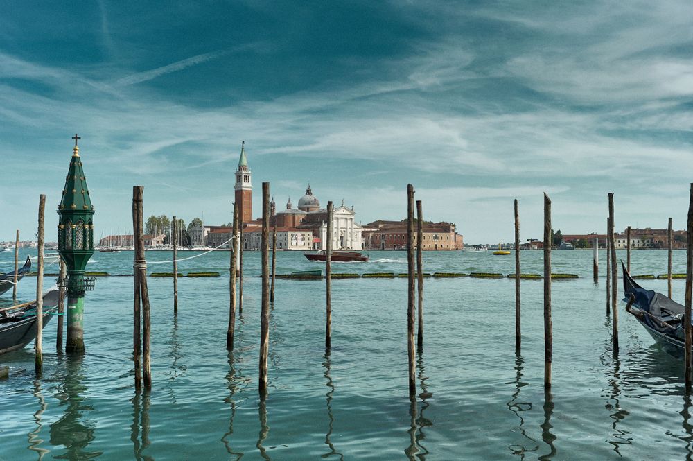 Mooring poles emerge from turquoise waters of Venice's Grand Canal with the city skyline in the distance.