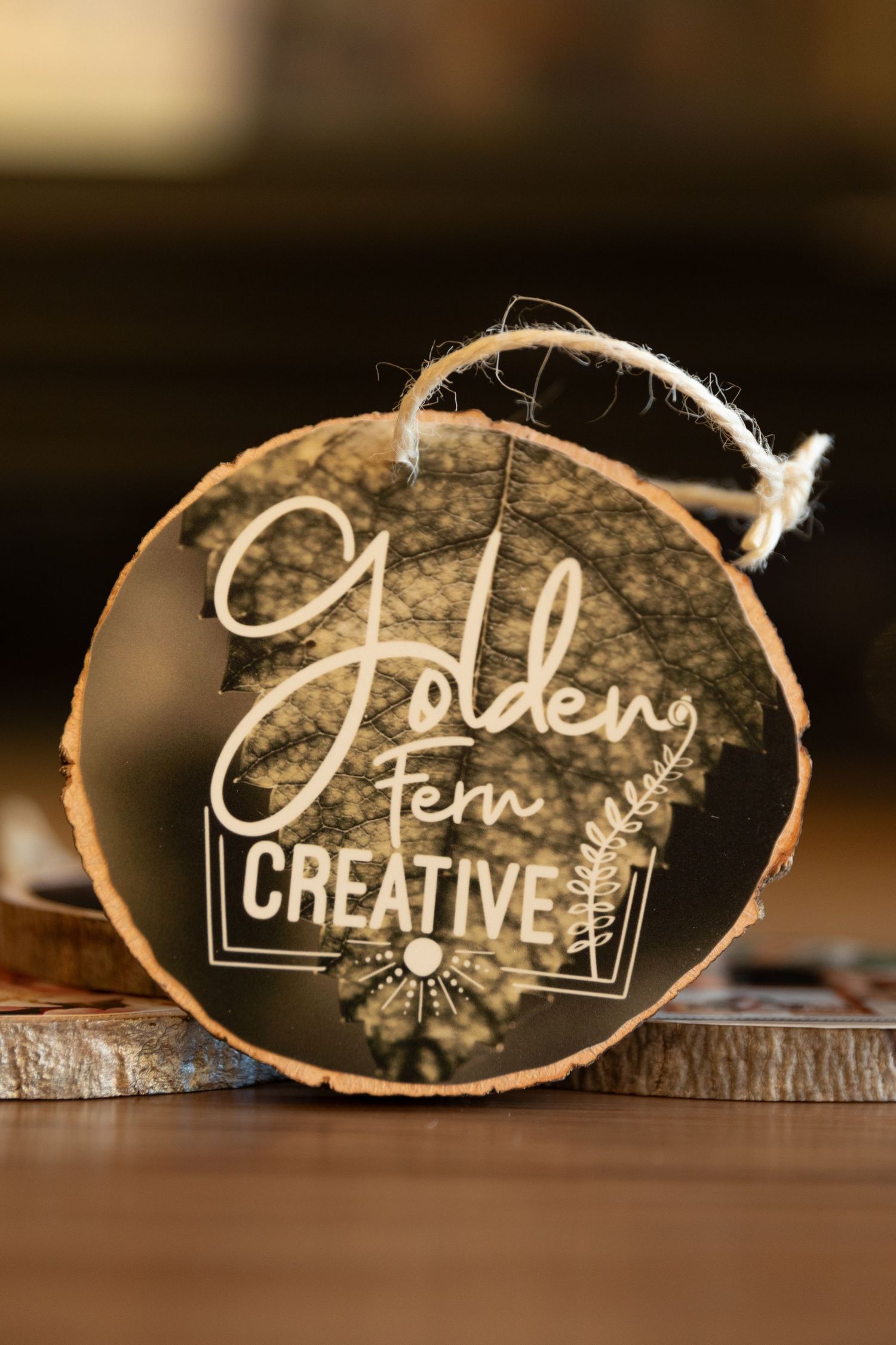 A rustic wooden slice ornament with 'Golden Fern Creative' logo and text etched in white against a dark background.