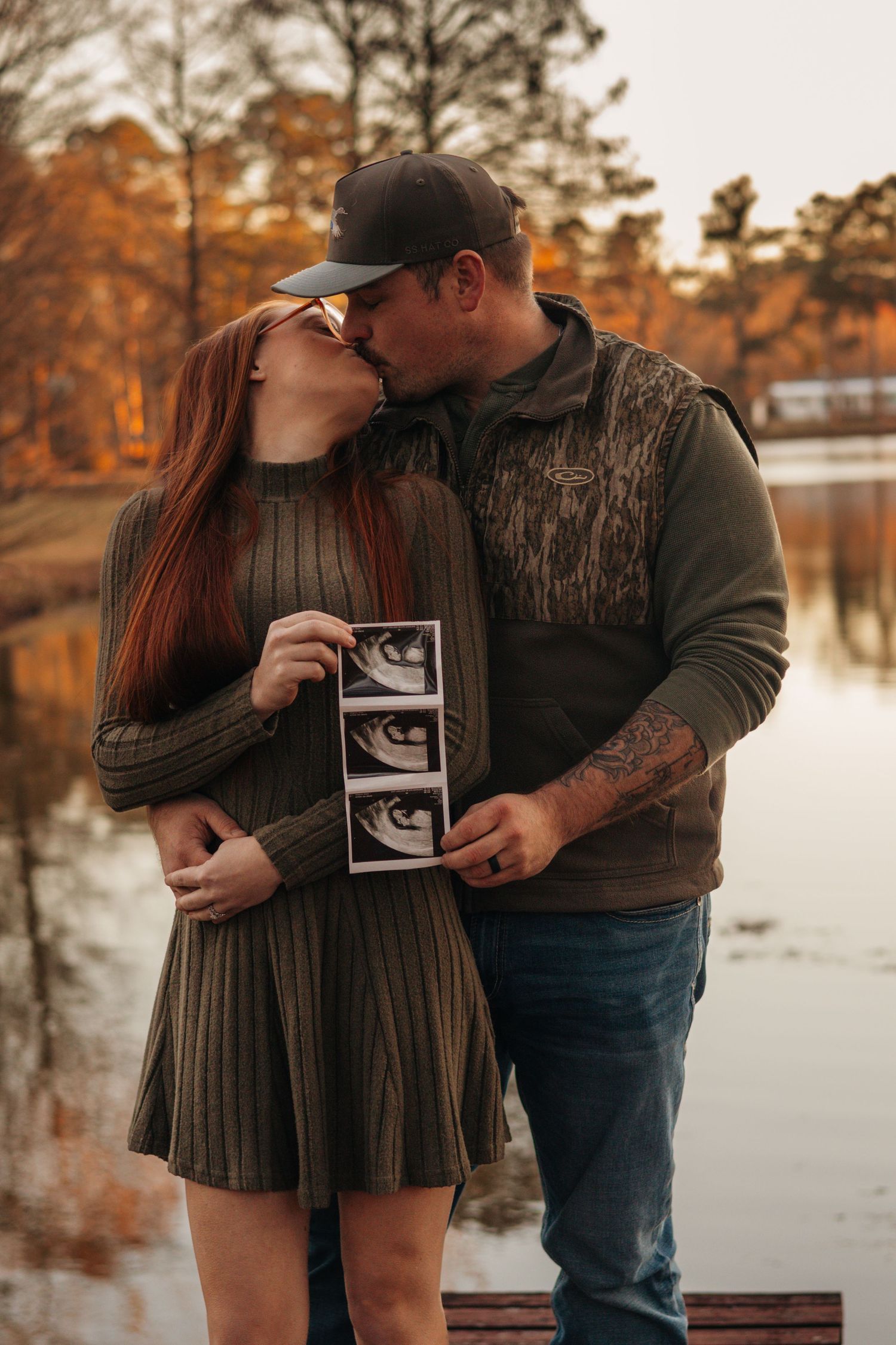 Expecting couple embracing beside a lake surrounded by autumn trees during a fall maternity photography session.