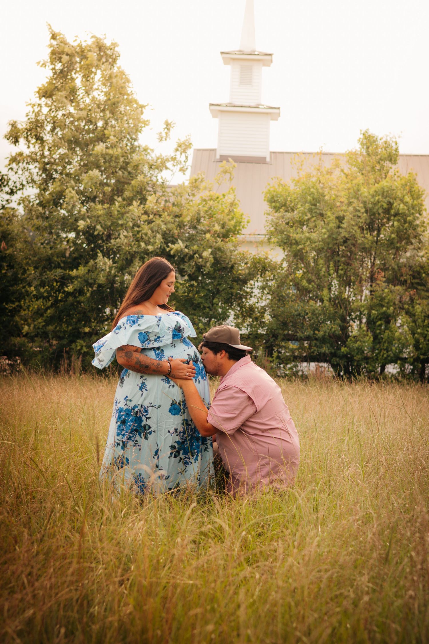Pregnant couple enjoying a maternity session together in a grassy field near a church at golden hour.