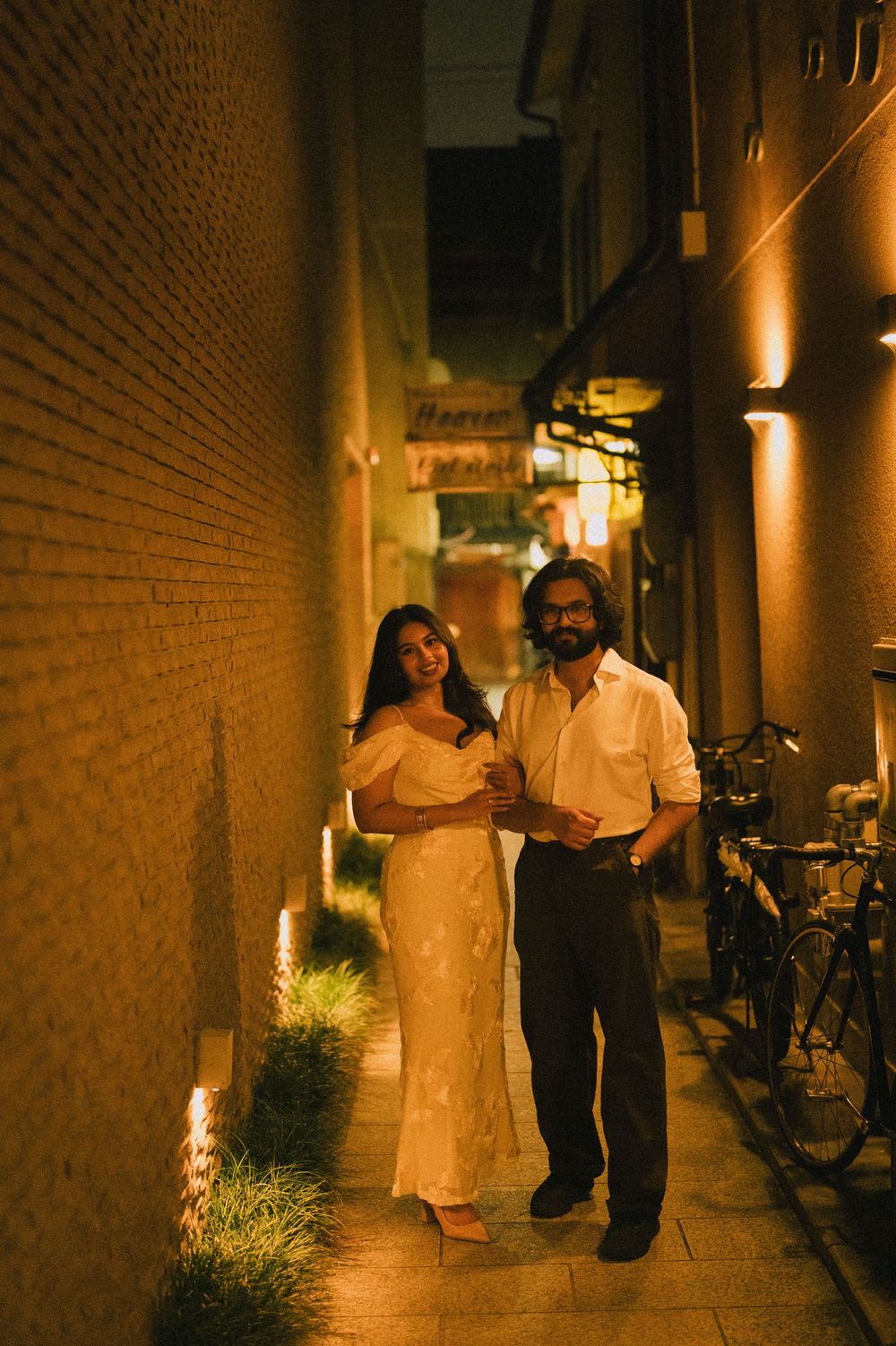 Couple posing in a warmly lit narrow alley at night, woman in white dress, man in white shirt.