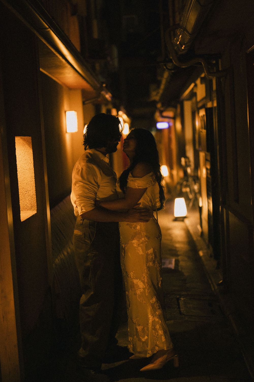 Couple embracing in a narrow warmly lit Japanese alley at night, golden lanterns glowing around them.