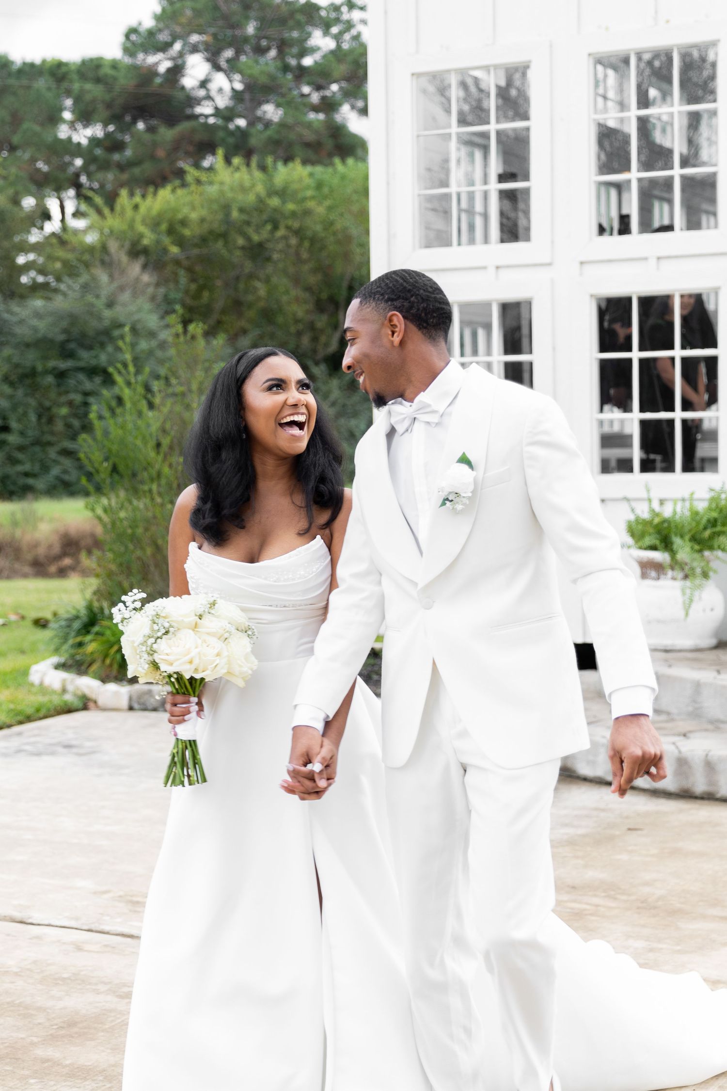 Joyful Black bride in white strapless gown and groom in white suit holding hands and laughing outside elegant white venue.