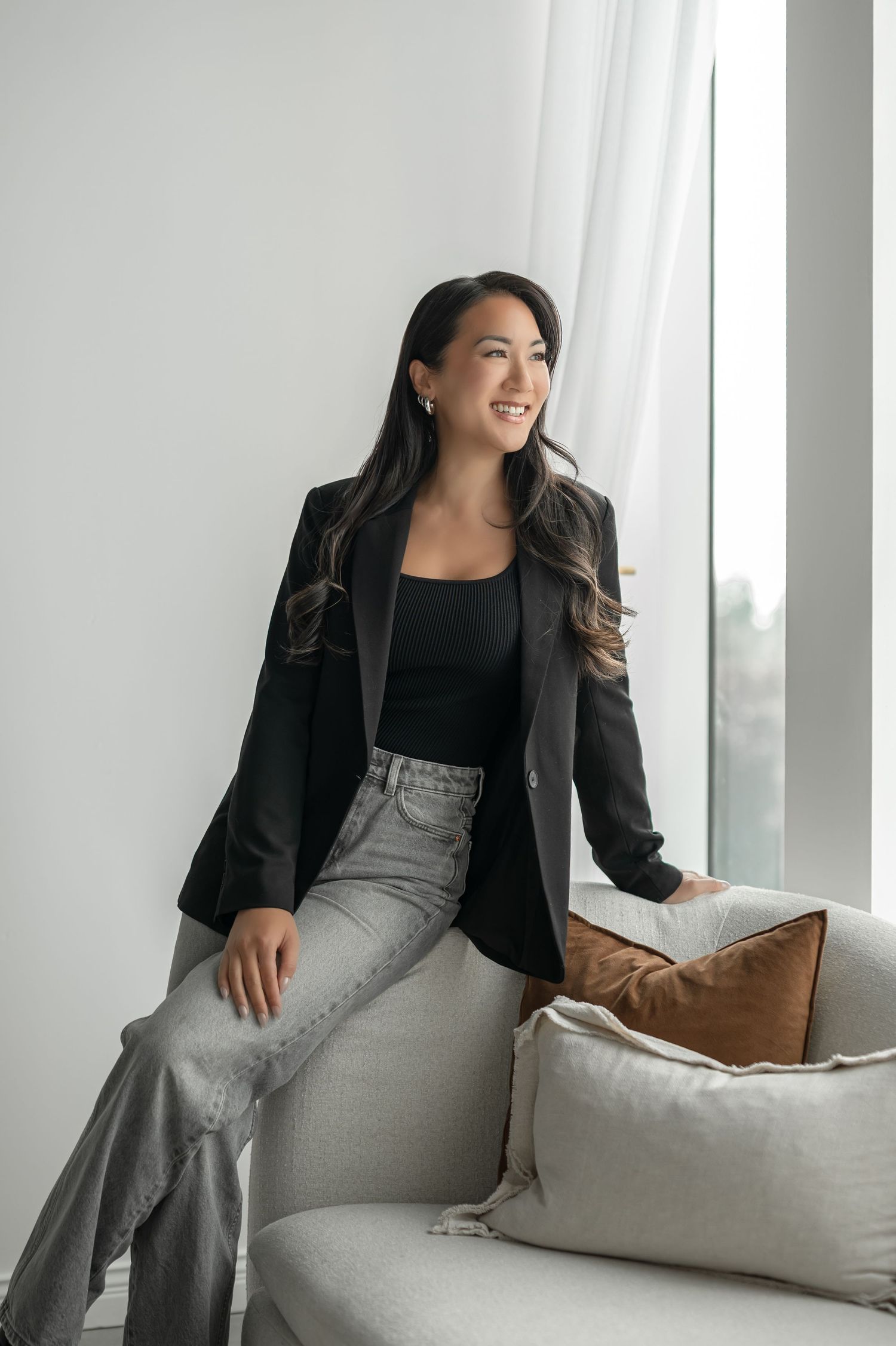 Professional woman in black blazer and gray pants relaxes on white couch near window in modern setting.