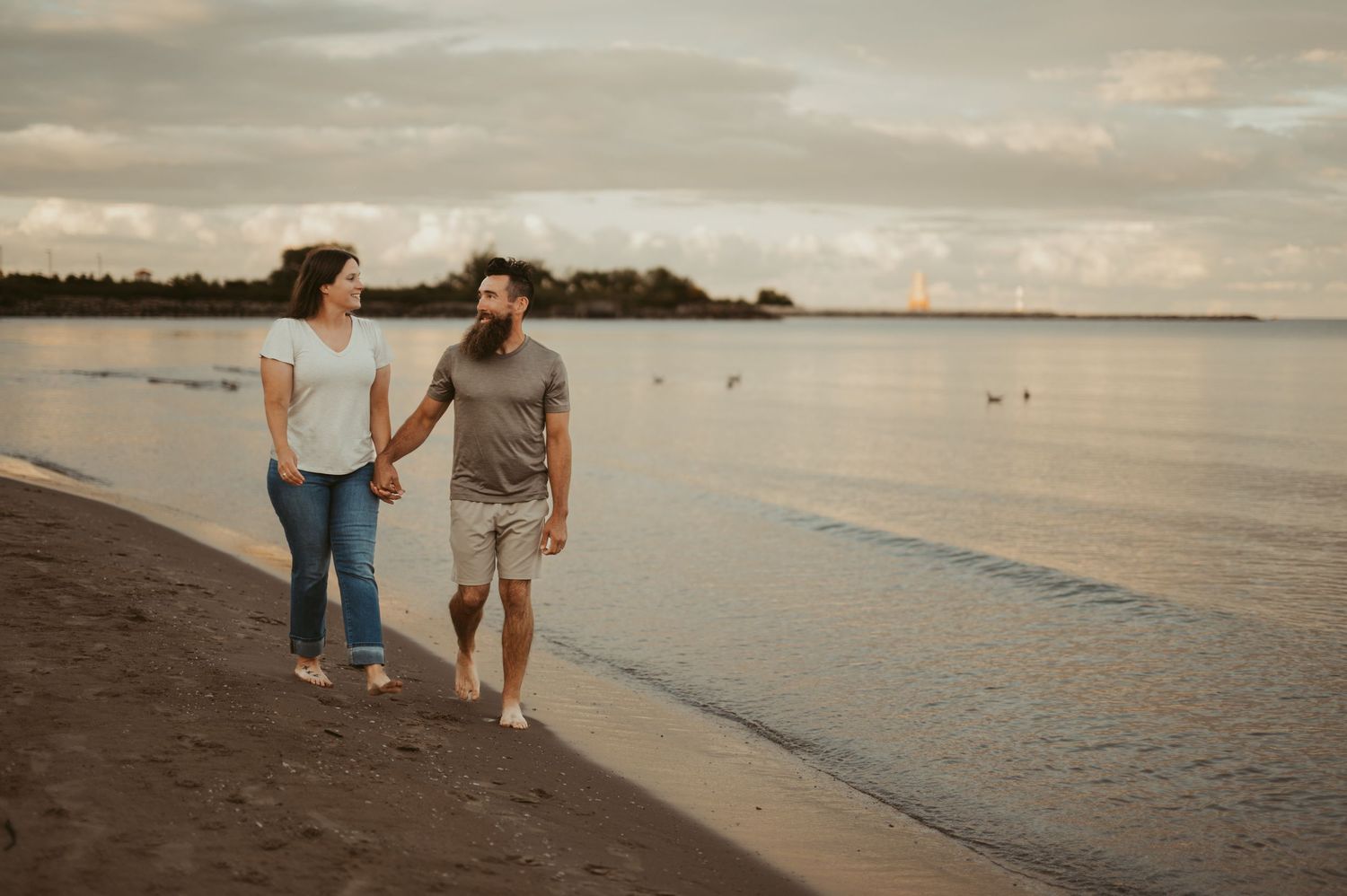 Couple holding hands walking along a sandy beach shore at sunset with calm water in the background.