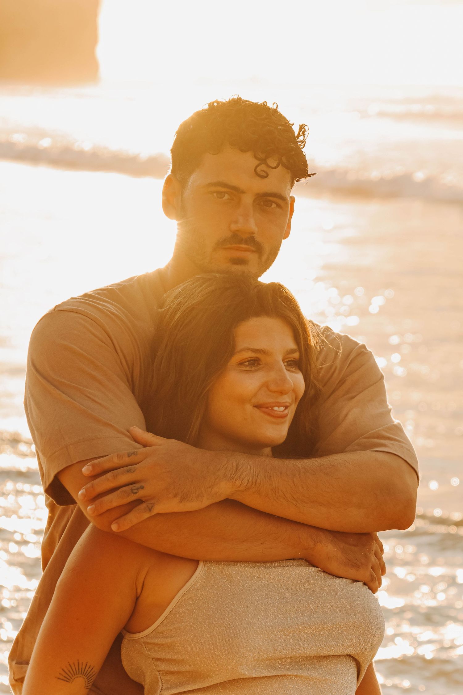 Man embracing woman from behind on beach at sunset, both bathed in warm golden light.