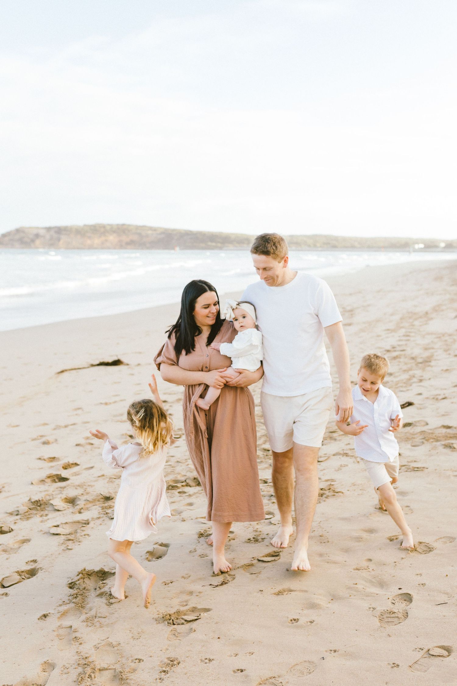 Family wearing neutral colors walks together on sandy beach during golden hour sunset photoshoot.