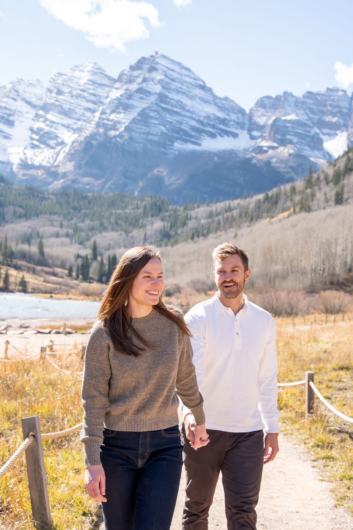 A couple walks hand in hand on a trail with snow-capped peaks of The Maroon Bells in the background