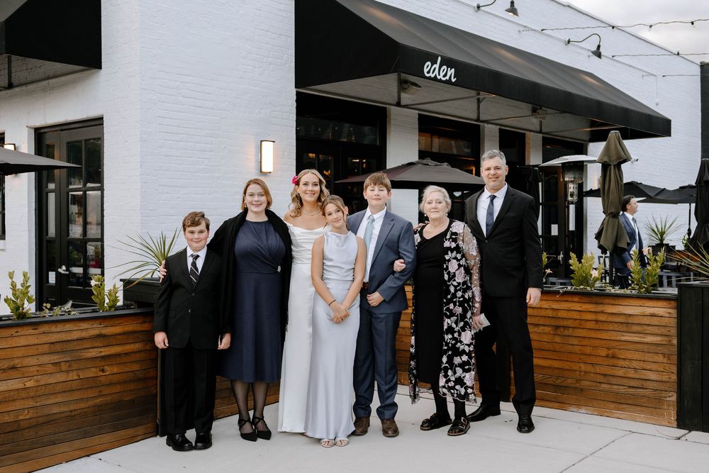 Wedding party stands outside a modern restaurant venue with wood paneling and white exterior walls.