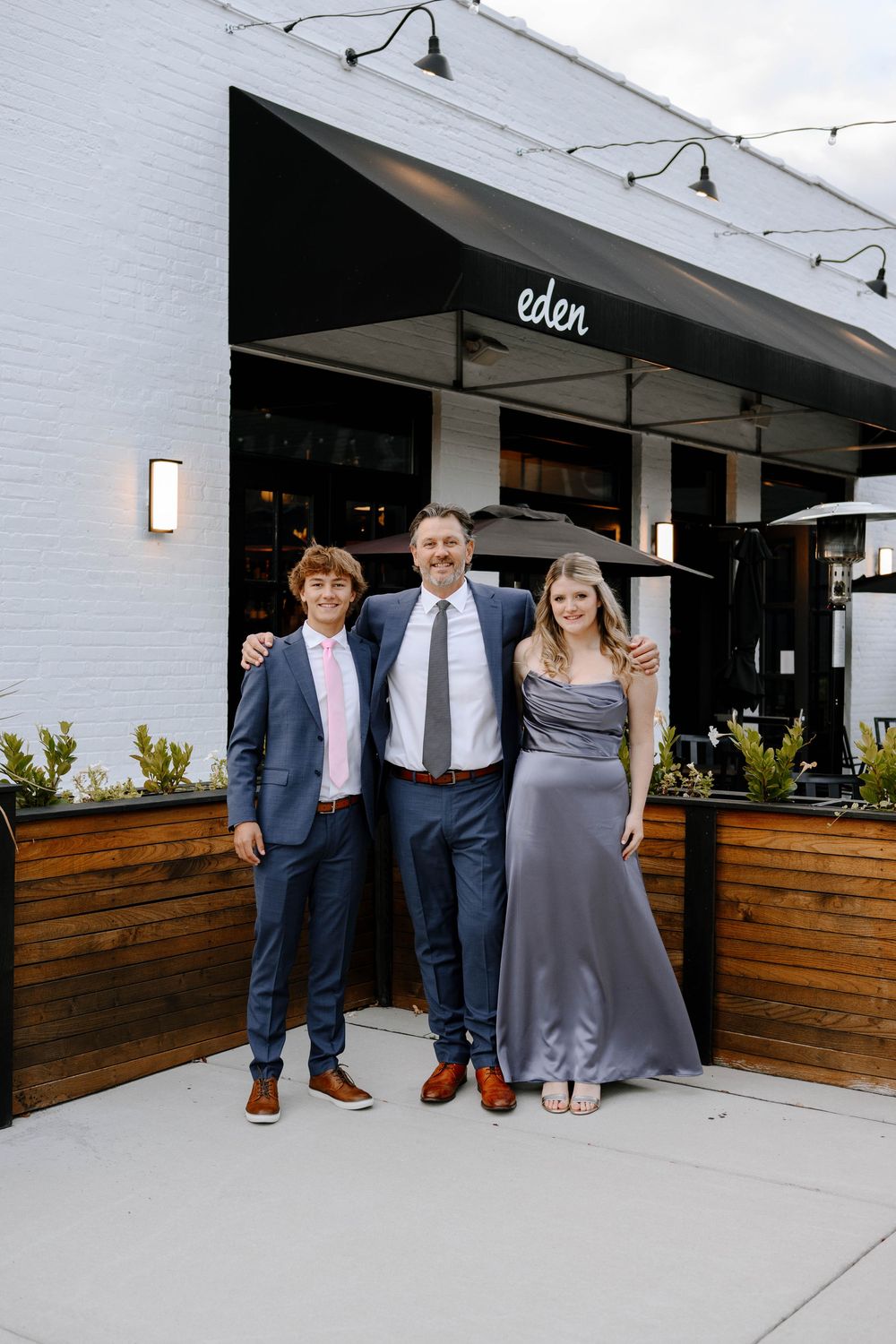 Wedding group gathers for photos outside modern venue with white brick walls.