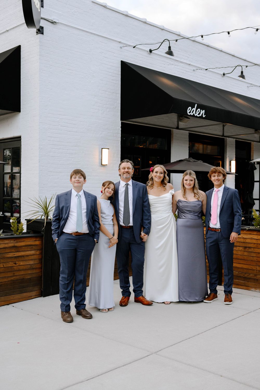 A wedding party poses outside a modern venue with white walls and wooden accents.