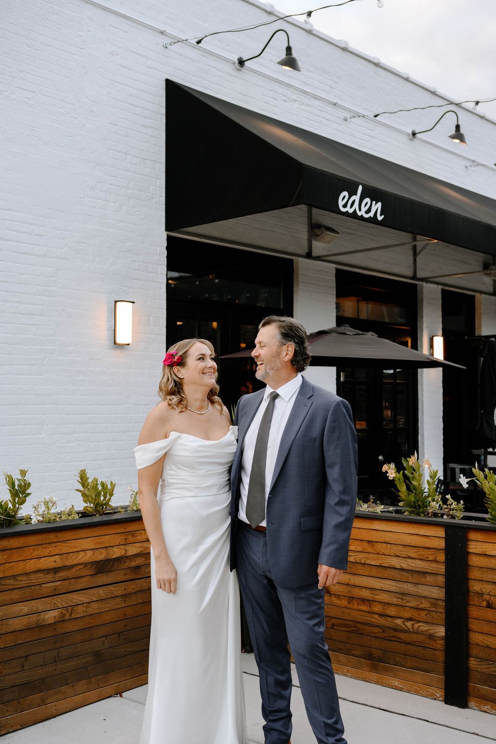 A couple shares a joyful moment outside Eden restaurant with white brick walls and wooden accents.