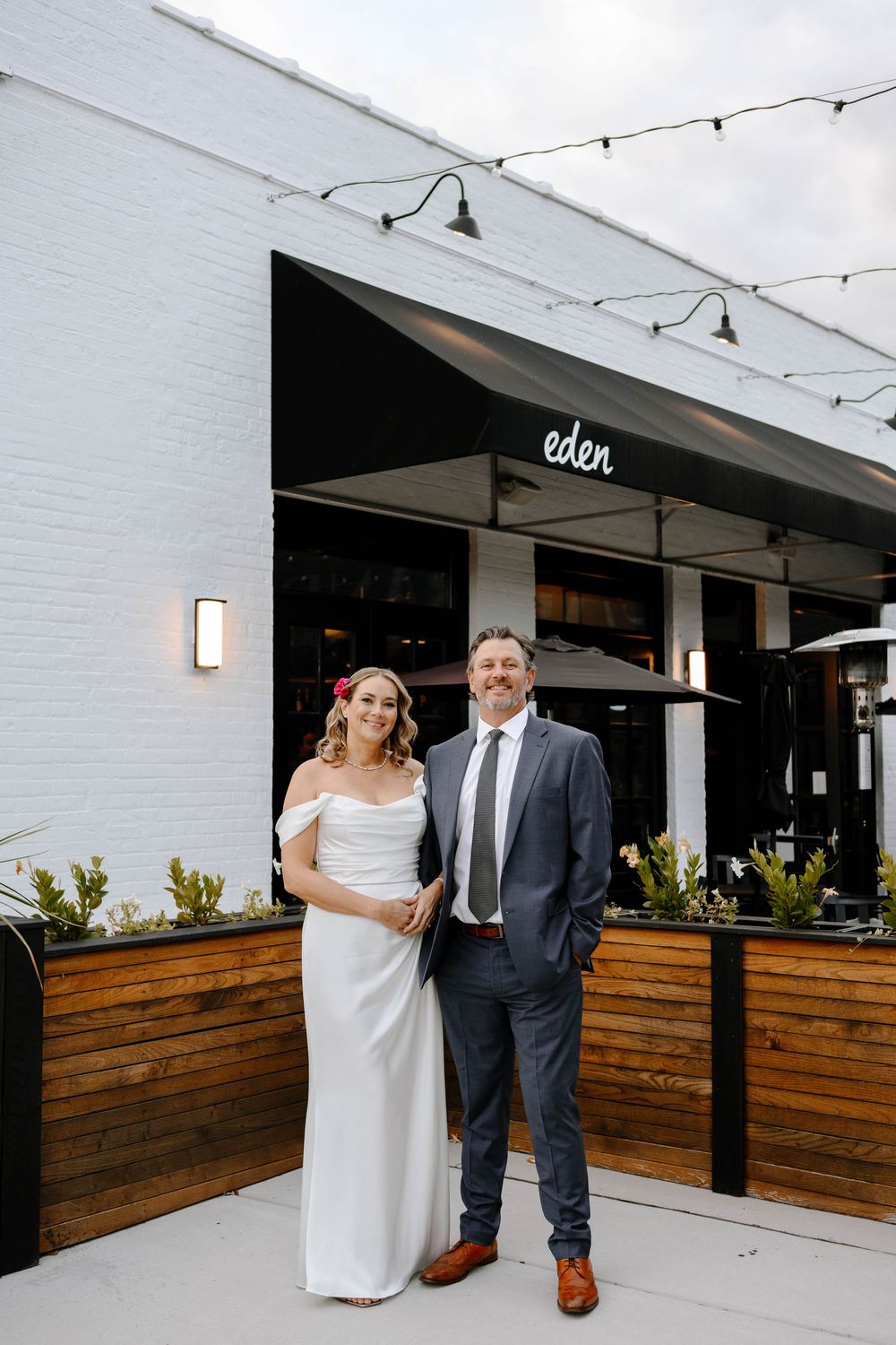 A couple poses outside a modern restaurant with wooden paneling and string lights at Eden.