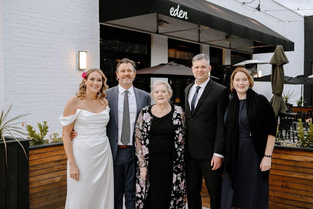 Wedding party group poses together outdoors at modern venue with wooden paneling and white walls.