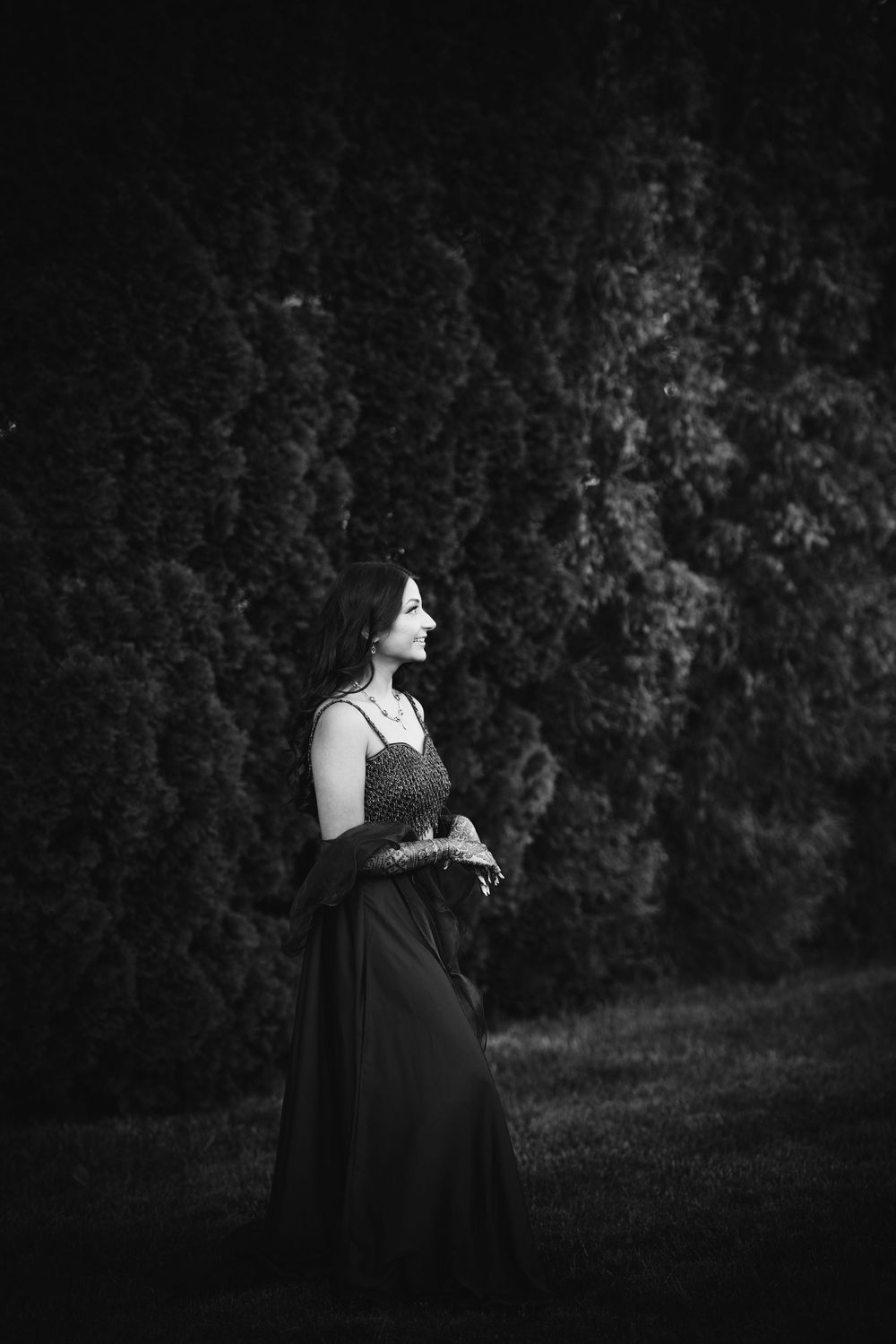 A dark and moody black and white portrait of a person in a dress standing against a shadowy natural backdrop.