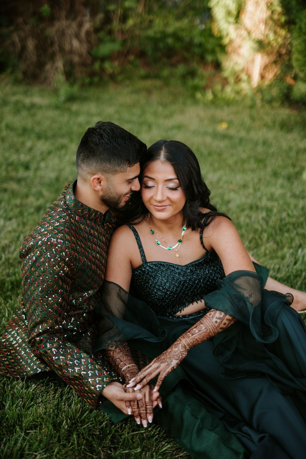 A couple in traditional South Asian attire share intimate moments during an outdoor photoshoot on grass.