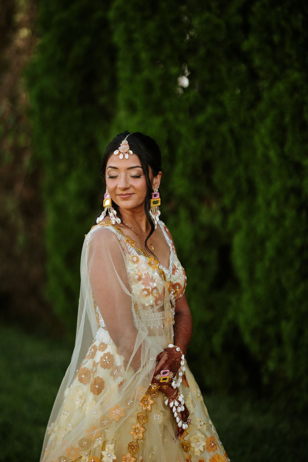 A bride in a sheer white and gold embroidered lehenga poses with closed eyes against a green outdoor background.
