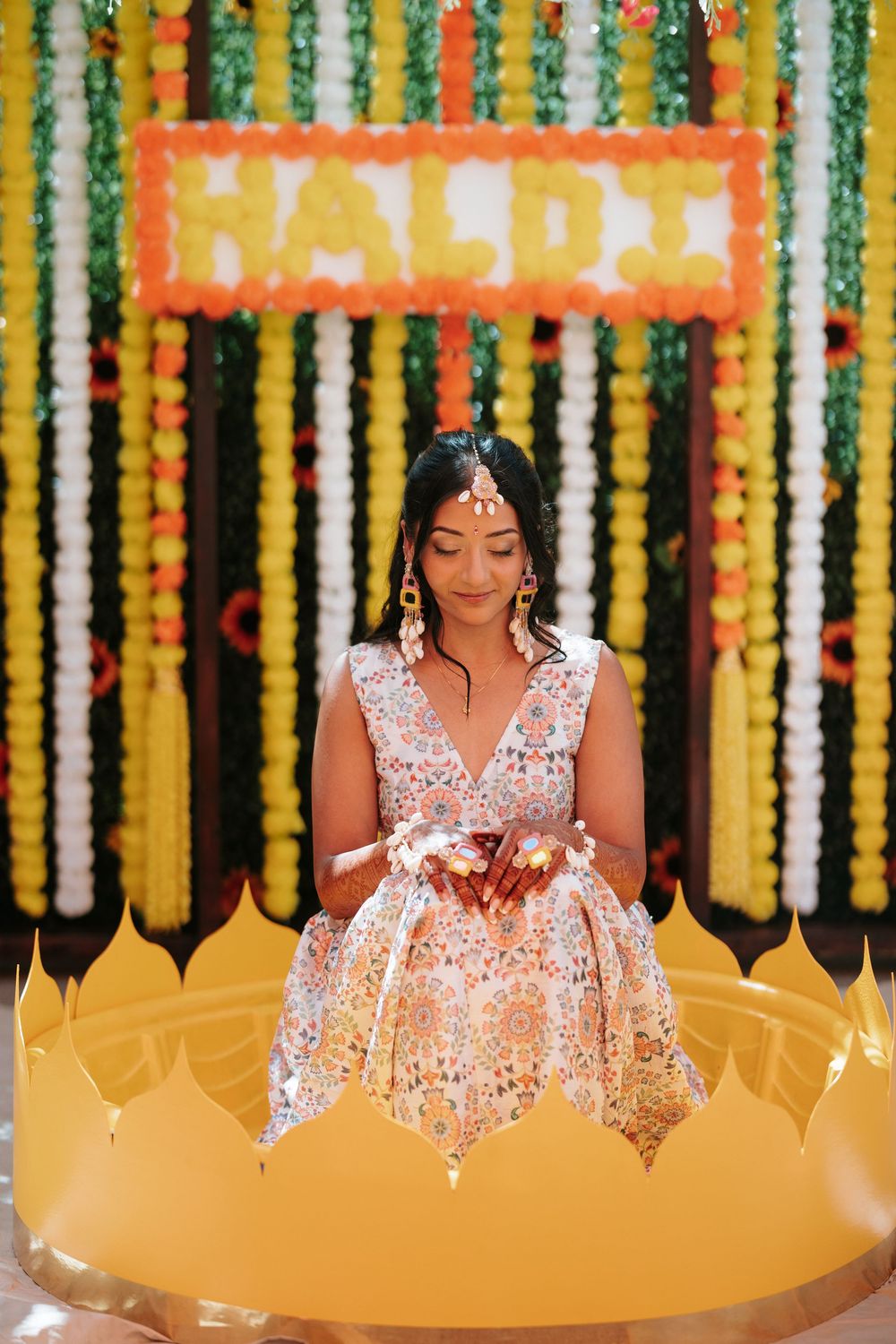 Woman in white lace dress poses against vibrant yellow and orange floral backdrop at traditional Indian celebration.