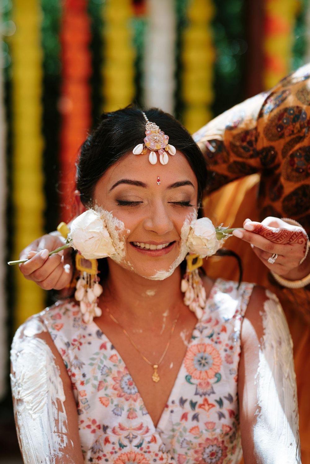 A traditional Indian bride smiles during the Haldi ceremony wearing a white outfit with floral embroidery and gold jewelry.