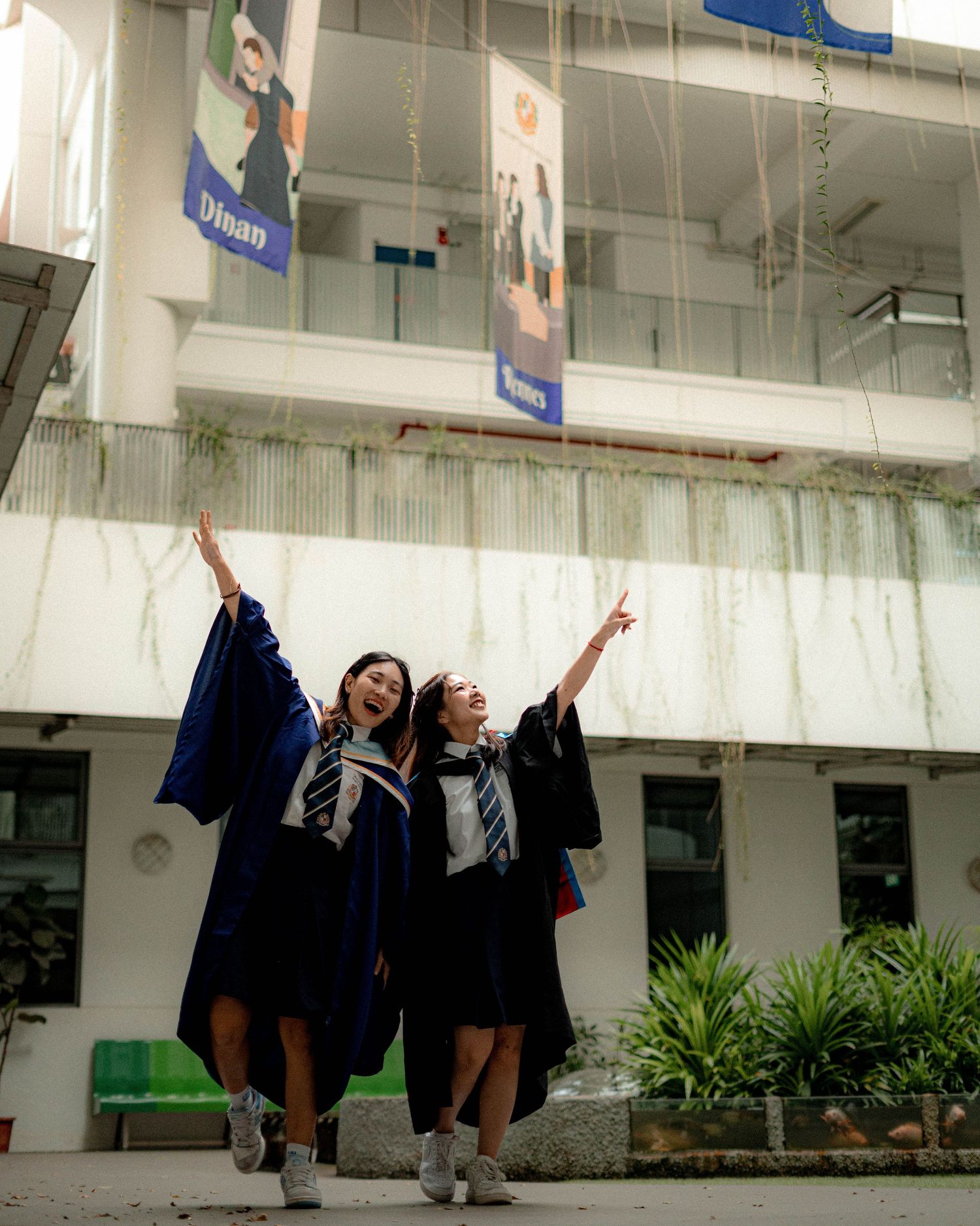 Excited graduates in blue gowns celebrate by raising their arms in front of a school building.