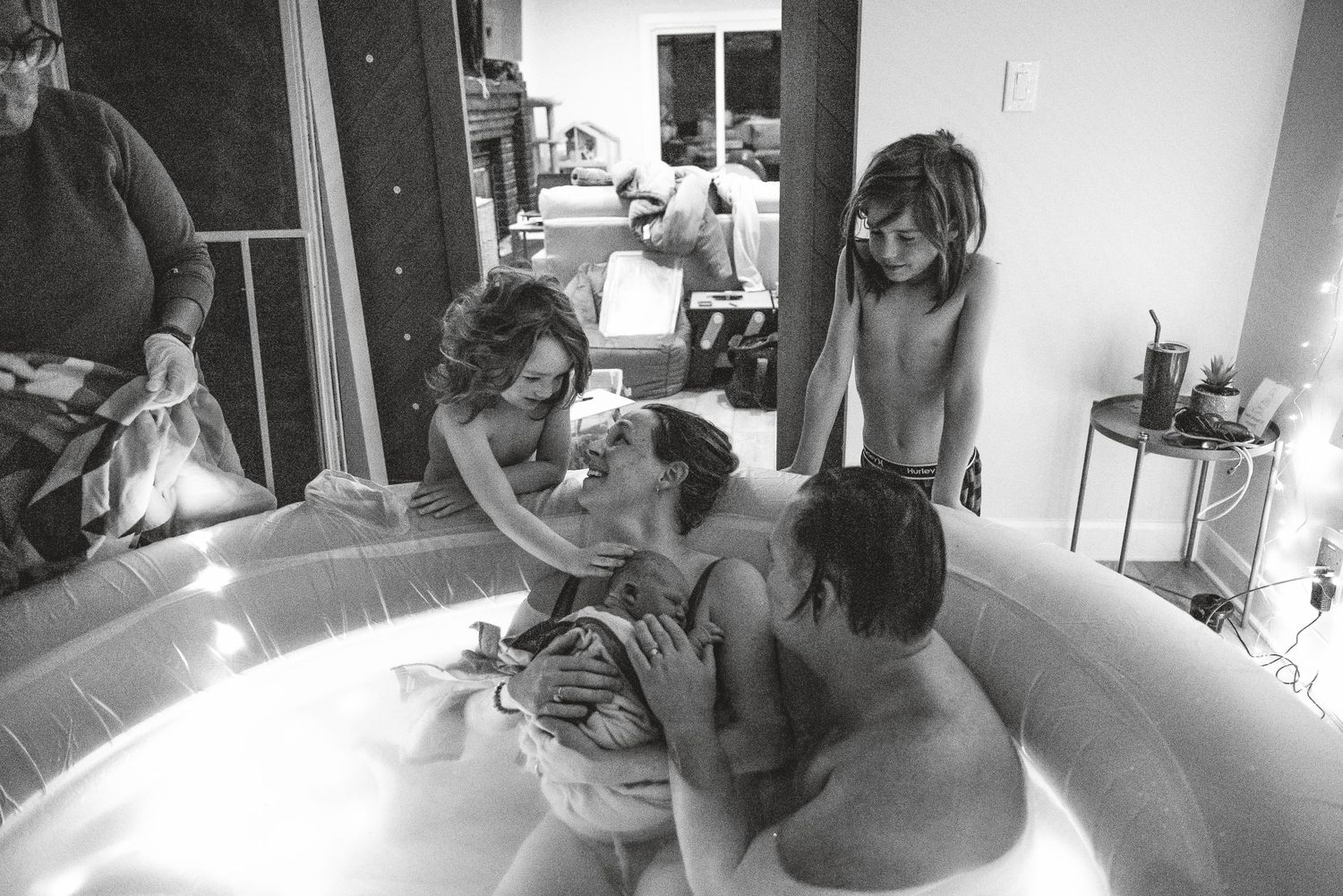 A black and white photograph shows people gathered around an inflatable birthing pool during a home water birth.