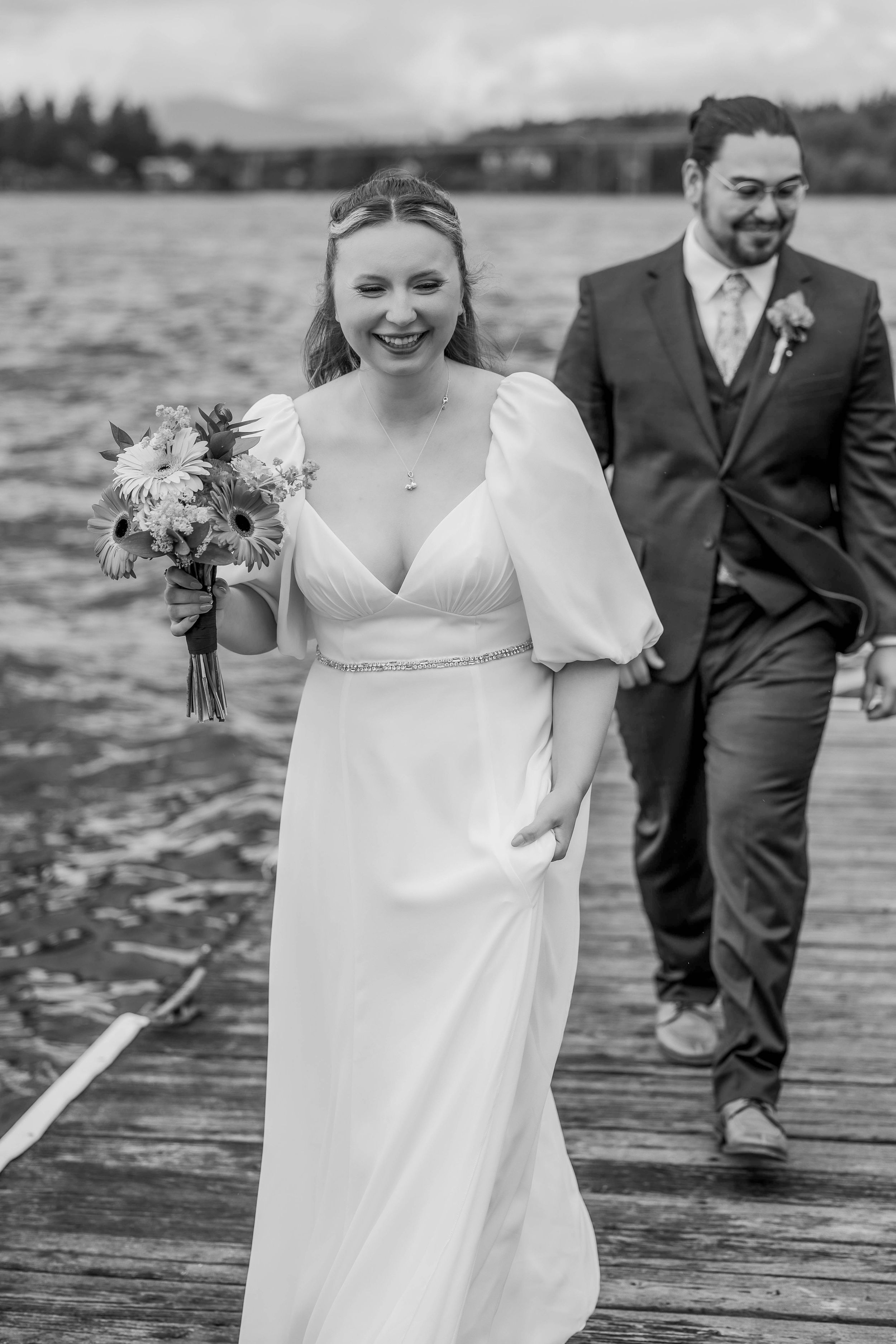 A joyful wedding couple walks along a wooden dock by a lake in black and white photography.