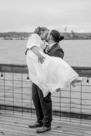 A romantic black and white wedding photo of a couple embracing on a waterfront dock with water and shoreline in background.