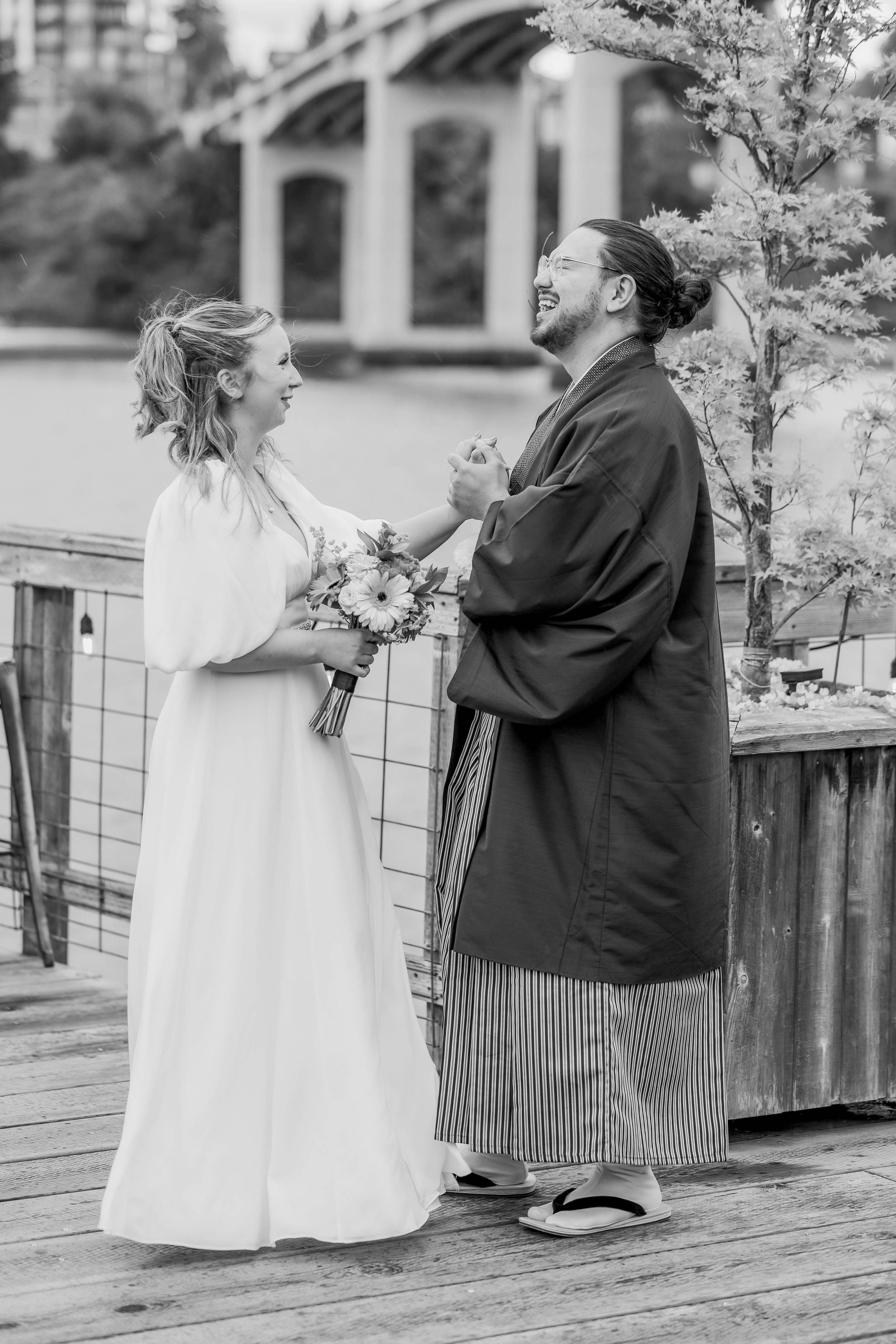 Black and white wedding photo showing a couple sharing a joyful moment on a wooden deck, bride holding a bouquet.