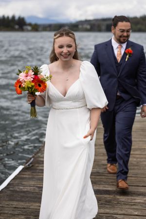A couple walks along a waterfront dock on their wedding day against a cloudy lake backdrop.