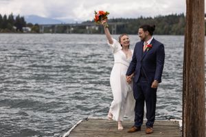 A pair in wedding attire stand joyfully on a wooden dock beside a scenic lake with mountains in the background.